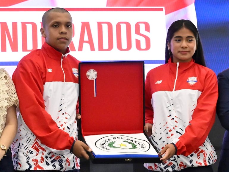 Sofía Martínez y Leonardo Acuña sostienen reconocimientos en ceremonia, rodeados de autoridades en ambiente festivo con decoraciones deportivas.