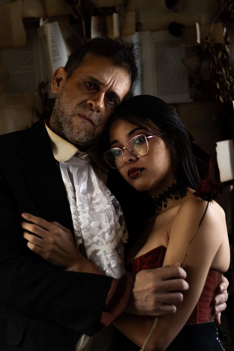 Hombre en traje oscuro y mujer en vestido rojo se abrazan, rodeados de decoraciones artísticas y sosteniendo una rosa negra.