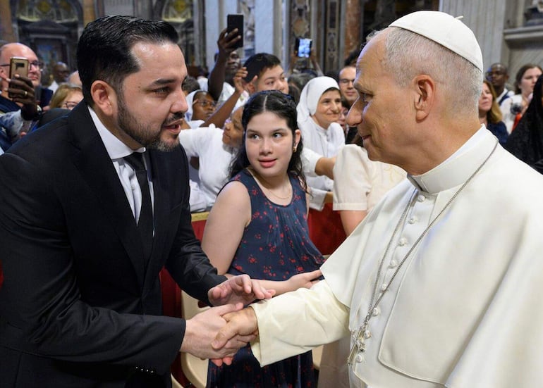 Luis Bello en traje negro estrecha la mano del papa León XIV, vestido de blanco, en un interior con ambiente religioso, rodeados de personas.