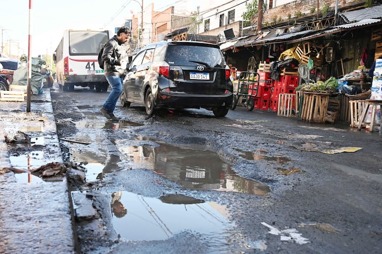 Hombre con chaqueta y jeans avanza por calle pavimentada con charcos, rodeado de cajas de frutas en el Mercado 4.