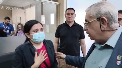 Mujer con suéter oscuro y camiseta roja gesticula mientras habla con hombre de gafas en sala de espera.