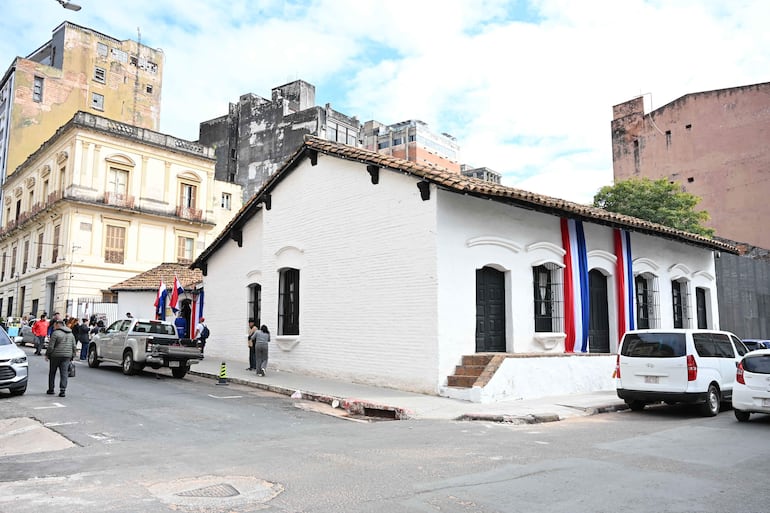 Fachada de casa colonial decorada con cintas de colores rojo, blanco y azul, vehículos estacionados en la calle.