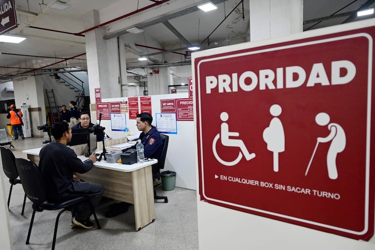 Hombres en oficina pública, uno asiste a otro en trámites, con cartel de prioridad visible en entorno de oficina.