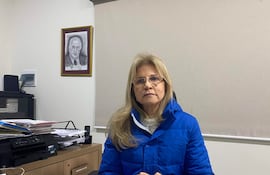 Mujer con cabello rubio lacio y gafas, vestida con chaqueta azul, sentada en un escritorio con documentos y una impresora.