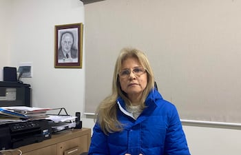 Mujer con cabello rubio lacio y gafas, vestida con chaqueta azul, sentada en un escritorio con documentos y una impresora.