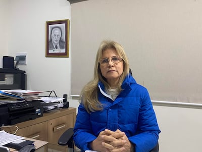 Mujer con cabello rubio lacio y gafas, vestida con chaqueta azul, sentada en un escritorio con documentos y una impresora.
