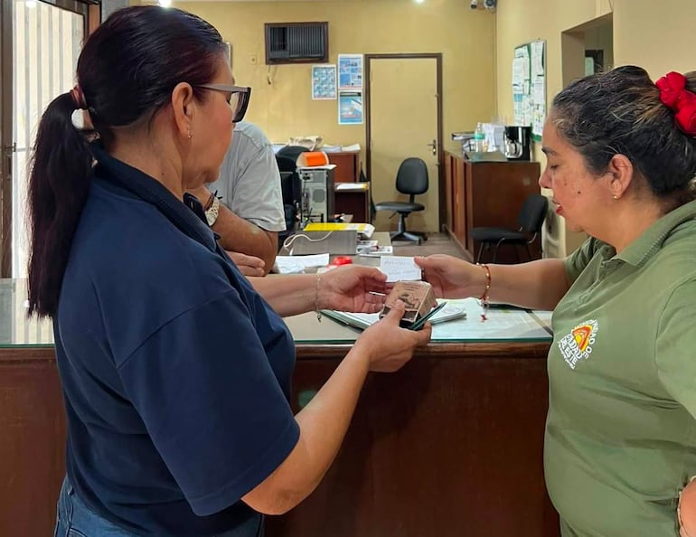 Dos mujeres en la oficina, una en coleta y otra con camiseta verde, intercambian documentos frente a un escritorio desordenado.