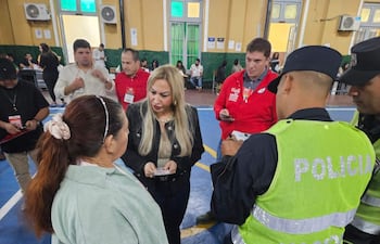 Dos mujeres conversan, una con chaqueta negra y otra en prenda verde, rodeadas de funcionarios y agentes policiales en un salón amplio.