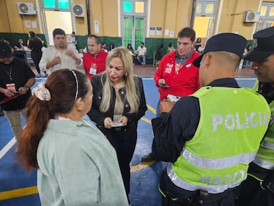 Dos mujeres conversan, una con chaqueta negra y otra en prenda verde, rodeadas de funcionarios y agentes policiales en un salón amplio.