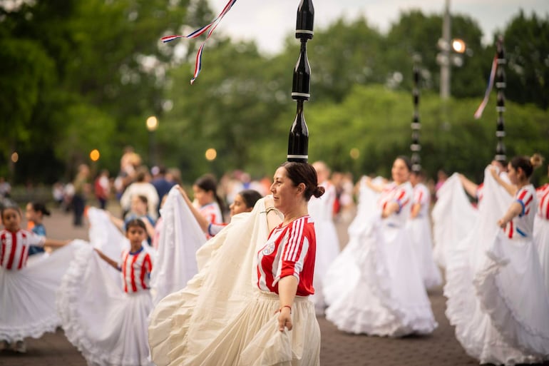 Mujeres bailan en grandes faldas blancas y camiseta roja, una lleva botellas en equilibrio sobre la cabeza en un evento cultural al aire libre.