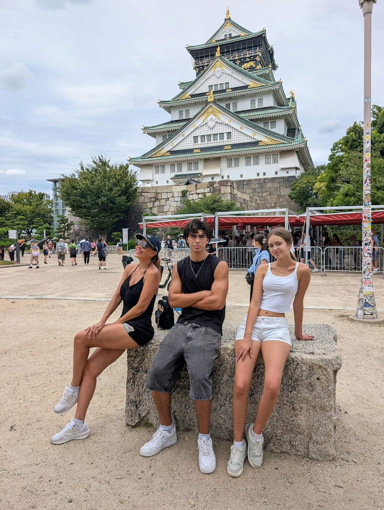 Mujeres en ropa colorida y un hombre en camiseta negra sonríen sentados sobre piedra, con un castillo japonés al fondo.
