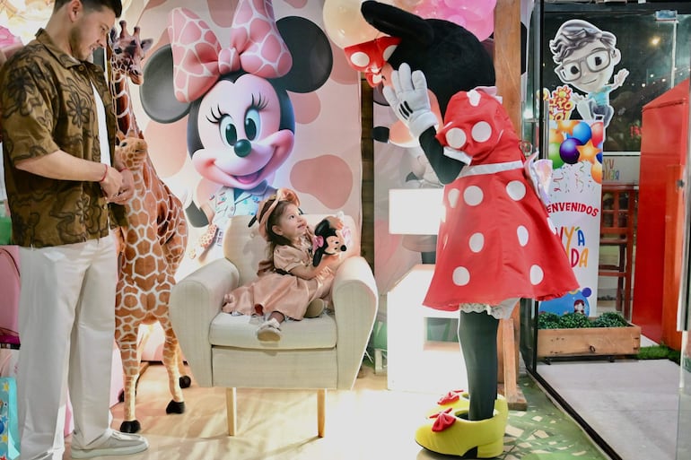 Ángel Romero sonríe, vestido con camisa floral, mientras una niña con vestido rosa saluda a Minnie Mouse en una fiesta infantil con globos.