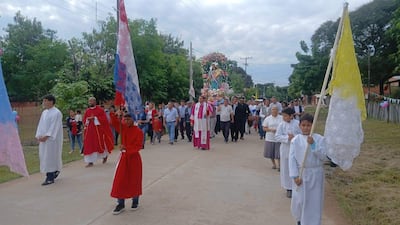 Tres figuras con túnicas religiosas, una en manto rojo, marchan con estandarte en celebración, con un entorno residencial y árboles.