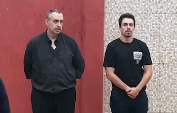 Fernando Barrionuevo en camisa y pantalón negro, Aitor Barrionuevo en camiseta negra, ambos con expresión seria en un entorno interior rojo.