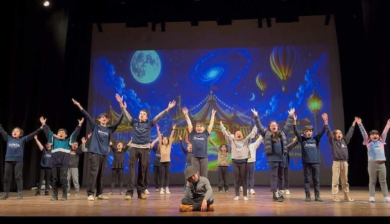 Grupo de jóvenes en escenario, con ropa oscura y camisetas coloridas, bailando bajo un telón con un cielo estrellado y globos aerostáticos.