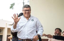 Hombre de pie con camisa blanca y gafas, sonriente, levanta mano derecha en un ambiente participativo con brazos de otros presentes.