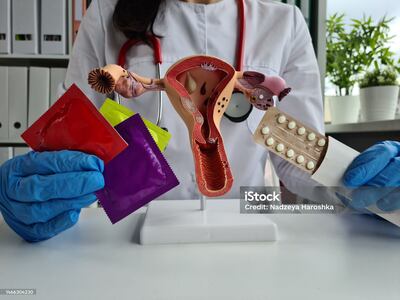 Una médica con guantes de látex sostiene preservativos, condones, pastillas anticonceptivas y muestra un útero de plástico