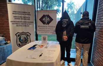 Dos hombres de espaldas en un entorno institucional oscuro, con documentos sobre una mesa y carteles visibles del Ministerio Público.