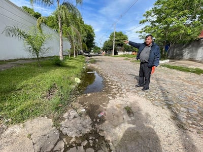 Hombre de chaqueta azul y pantalones oscuros, señalando, con un documento rojo en la mano, en una calle adoquinada.