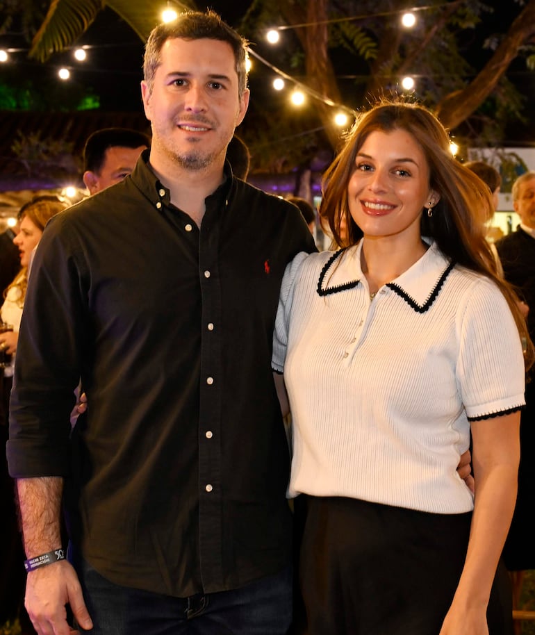 Mujer con blusa blanca y detalles negros sonríe junto a hombre de camisa negra en un ambiente festivo con luces colgantes.