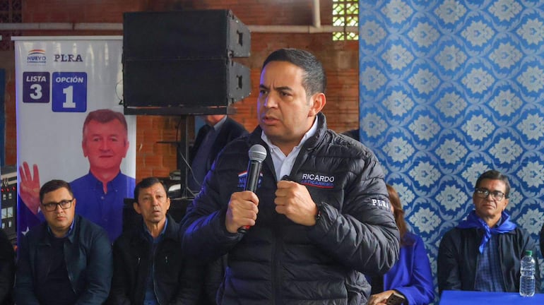 Ricardo Estigarribia en abrigo negro con micrófono, habla ante una multitud en ambiente con decoración azul y un cartel de 'PLRA'.