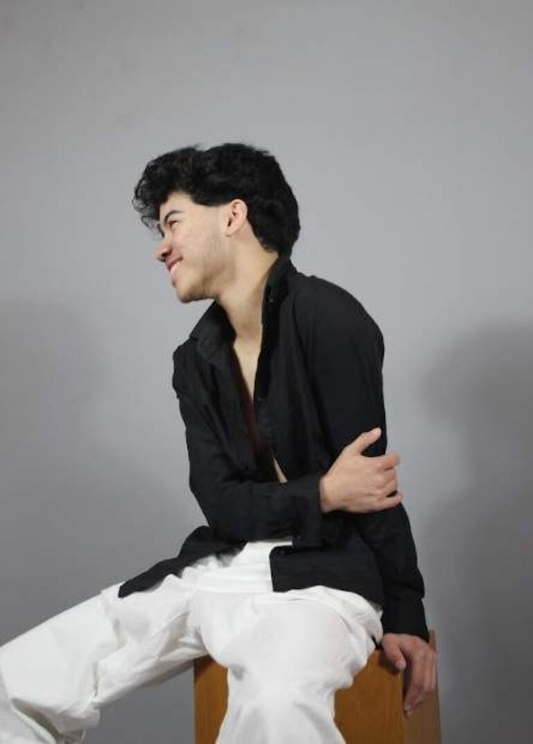 Joven en camisa negra y pantalones blancos, sonríe sentado sobre una silla en un fondo gris neutro.