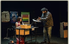 Mujer con cabello largo y camisa blanca escucha a hombre de chaqueta oscura y jeans en un auditorio con carteles informativos.