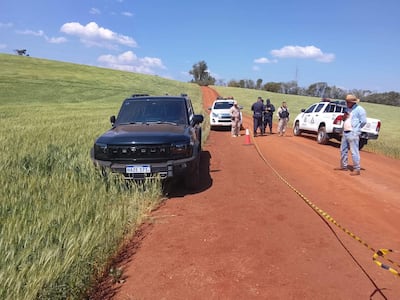 Cuatro personas, incluyendo dos uniformados de policía, en un camino rural con vehículos y cinta de señalización.
