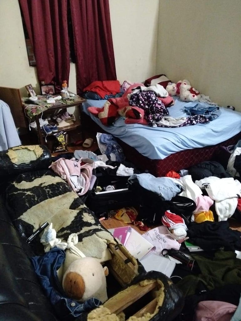 Interior desordenado de un dormitorio con camas deshechas, ropa esparcida y un sofá negro desgastado.
