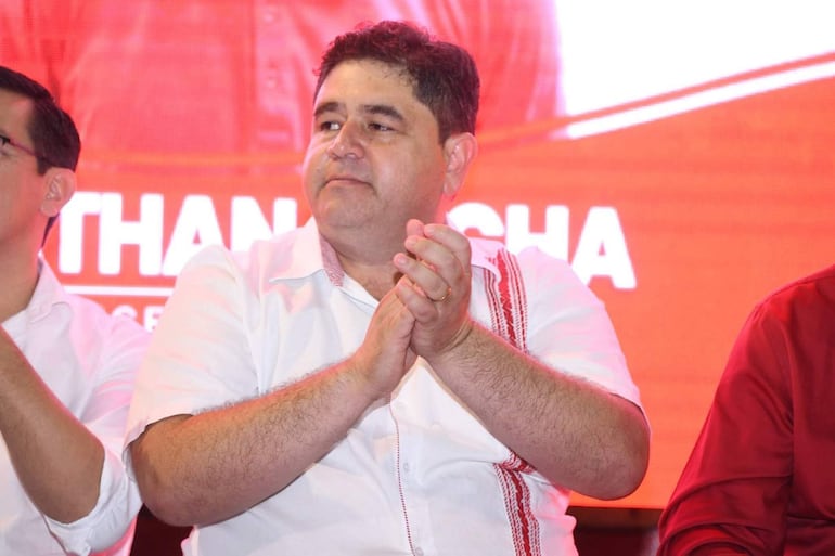 Hombre aplaudiendo con camisa blanca y detalles rojos, mientras otros dos hombres lo escuchan en escenario con fondo rojo.