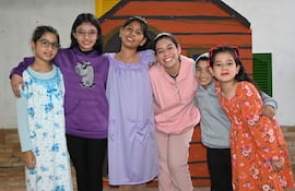 Grupo de seis niños y una joven adulta sonriendo, vestidos con ropa colorida en un entorno decorado con temática de casa.