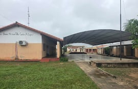 Escuela básica de un piso con fachada beige y naranja, césped verde, sin personas visibles y un perro negro en el entorno.