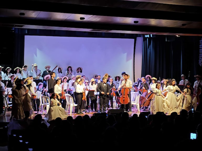 Músico en vestimenta blanca tocando violonchelo, acompañado por bailarinas con vestidos blancos y una orquesta activa al fondo.