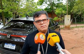 Monseñor Osmar López vestido con atuendo religioso, rodeado de árboles mientras es entrevistado. Micrófonos de medios apuntando hacia él.