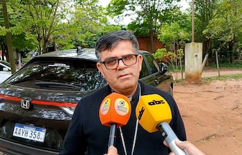 Monseñor Osmar López vestido con atuendo religioso, rodeado de árboles mientras es entrevistado. Micrófonos de medios apuntando hacia él.