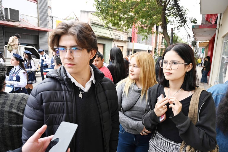 Tres jóvenes, un hombre con abrigo negro y dos mujeres, conversan en un ambiente festivo con banda al fondo.
