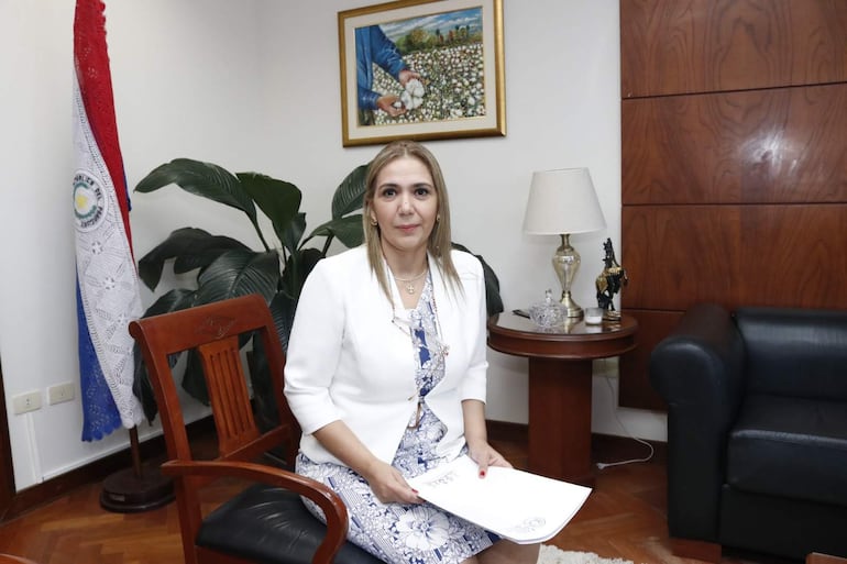 Nancy Salomón, mujer con cabello largo y blusa clara, sostiene documentos en un ambiente formal decorado con plantas y una pintura.