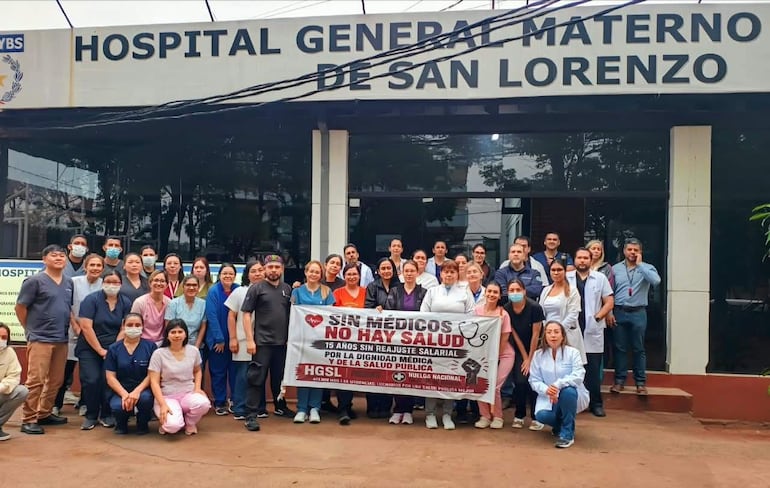 Médicos y personal de salud en uniformes diversos, sosteniendo pancarta con mensaje de protesta frente a un hospital.