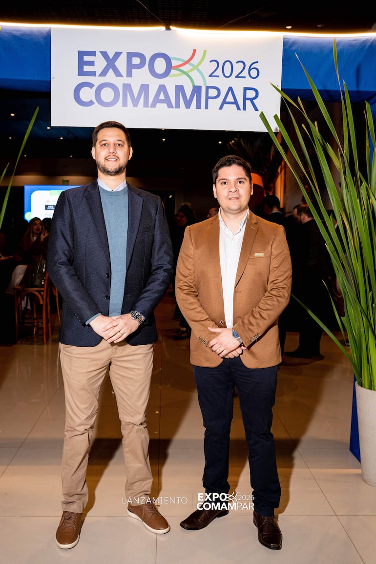 Dos hombres en traje oscuro y abrigo marrón, serios, en un evento con letrero de EXPO COMAMPAR 2026 en el fondo.