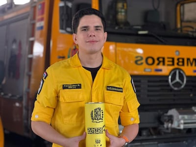 Bombero joven en uniforme amarillo, sosteniendo un cartel con texto y número telefónico, frente a un camión de bomberos amarilllo.