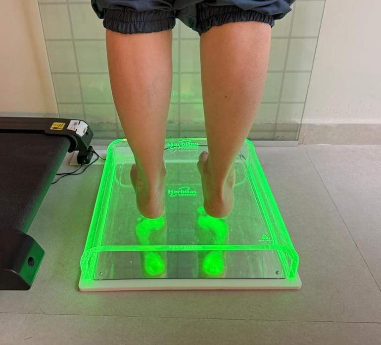 Persona de pie, con pantalones cortos deportivos, en plataforma iluminada con luz verde en un ambiente clínico.