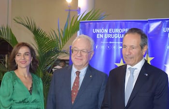 Tres hombres en un evento formal, sonrientes; mujer con vestido verde y hombres en trajes, fondo de pancarta que dice 'UNIÓN EUROPEA EN URUGUAY'.