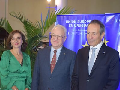 Tres hombres en un evento formal, sonrientes; mujer con vestido verde y hombres en trajes, fondo de pancarta que dice 'UNIÓN EUROPEA EN URUGUAY'.