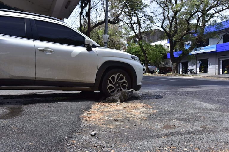 Vehículo blanco estacionado junto a un bache que emite agua, con edificios y árboles de fondo en un día soleado.