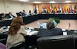 Varias personas vestidas de forma formal en una mesa rectangular con plantas y banderas de países al fondo, algunos tomando notas.