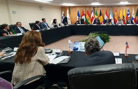 Varias personas vestidas de forma formal en una mesa rectangular con plantas y banderas de países al fondo, algunos tomando notas.