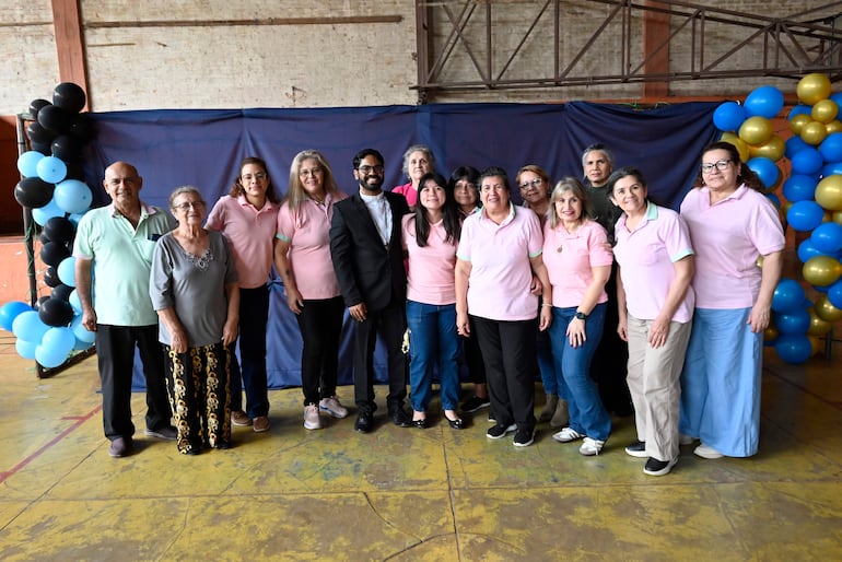 Catorce personas sonrientes posan en un fondo decorado con globos azul y negro, algunos llevan camisetas rosas.