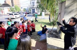 Grupo de niños con camisetas rojas escuchan atentamente a dos educadores, uno de ellos sentado en el suelo y el otro de pie en un entorno urbano.
