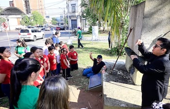 Grupo de niños con camisetas rojas escuchan atentamente a dos educadores, uno de ellos sentado en el suelo y el otro de pie en un entorno urbano.