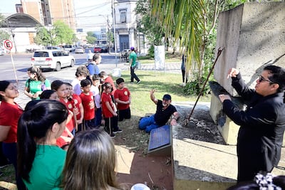 Grupo de niños con camisetas rojas escuchan atentamente a dos educadores, uno de ellos sentado en el suelo y el otro de pie en un entorno urbano.